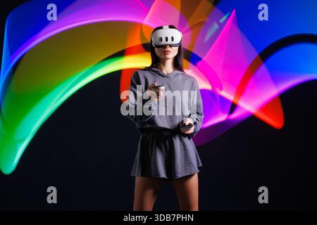 Young woman in VR headset playing virtual reality game with motion controllers, neon light trails in futuristic immersive environment. Stock Photo