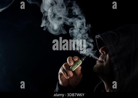 Mysterious hooded man vaping with e cigarette, blowing dense smoke clouds on black background, modern lifestyle and health risk. Stock Photo