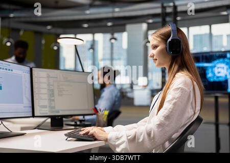 Young female programmer at work training machine learning models for anomaly detection automation. Tech startup employee wearing headphones, listening music and using artificial intelligence Stock Photo