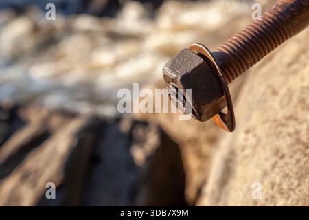 Macro view of an old rusty metal bolt with nut and washer fixed in rock, with a soft blurred river stream in the background. Stock Photo