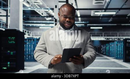 Smiling admin using tablet in in data center maintaining infrastructure for workloads. Joyful IT professional in server farm utilizing high tech server clusters for parallel processing, camera A Stock Photo