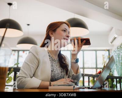 Young professional woman uses smartphone while working on a laptop at a cafe table. Multitasking and modern business communication concept. Stock Photo