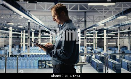 Jolly IT professional monitors server room activity using tablet in facility. Happy data center technician reviews rigs tests results with device to ensure systems are working correctly, camera A Stock Photo