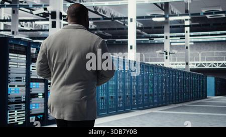 Admin walking in data center, doing inspection on infrastructure tech handling user requests. IT professional in server farm analyzing traffic patterns of equipment components, camera A Stock Photo