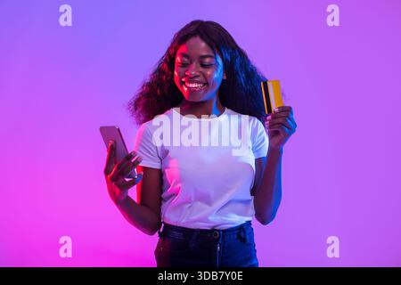 Cheerful Young Woman Engages in Online Trading With Smartphone and Credit Card in Vibrant Neon Light Stock Photo