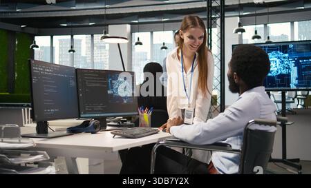 Young engineer and coworker with paraplegia doing brainstorming in office, reviewing AI system metrics. Junior dev and colleague in wheelchair talking, tuning neural networks using PC, camera A Stock Photo