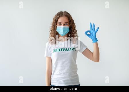 Young Volunteer Woman in Medical Mask and Gloves Making OK Gesture Against Light Wall Stock Photo