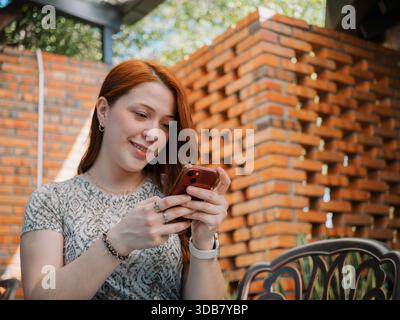 A young woman smiles while texting on her smartphone in an outdoor seating area with a brick wall background. Stock Photo