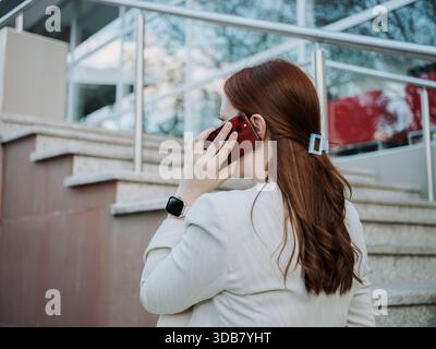 Businesswoman seen from behind using a smartphone while walking near an office area. Urban commute and connectivity concept. Stock Photo