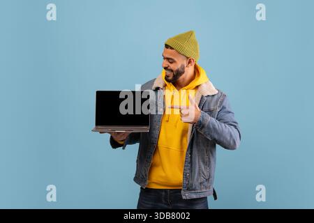 Man Holds a Laptop and Smiles While Wearing a Yellow Hoodie and Denim Jacket Against a Blue Background Stock Photo