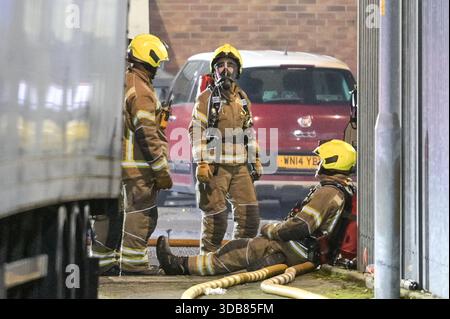 Newfield Close, Bloxwich 14th December 2025 - A firefighter rests after fighting the fire and another kits up ready to continue to help contain the inferno. - Ten West Midlands Fire Service appliances have been sent to a large fire in a Bloxwich Food Factory on Sunday evening. Initial 999 calls came in reporting smoke in the area near Walsall. The fire has largely effected one business, ‘G Simmons and Sons Pork Scratchings, Crackling and Crunch'. Fire engines surround the building with long hose relays stretching several hundreds of feet. Credit: British News and Media/Alamy Live News Stock Photo