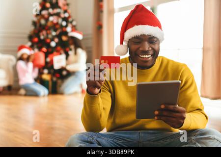 Man in Red Hat Using Tablet for Online Shopping During Holiday Season With Family Nearby Stock Photo