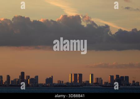 The Miami skyline is silhouetted against dramatic clouds and warm evening light as the sun sets over Biscayne Bay. Stock Photo