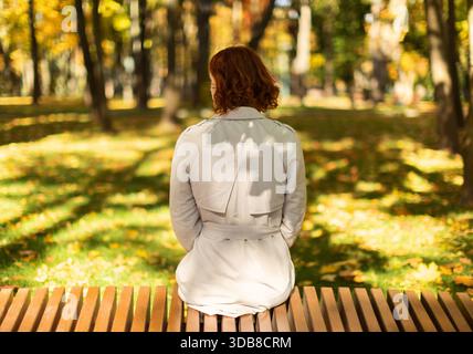 Woman Sits on a Bench in a Park Surrounded by Autumn Leaves on a Sunny Day Stock Photo