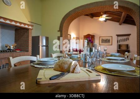 A warm, inviting dining room scene featuring a long wooden table set for a meal, fresh bread, wine glasses, and a brick archway Stock Photo