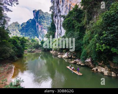 Two people glide peacefully across the calm river on a bamboo raft in Khao Sok, Thailand. Towering limestone cliffs and dense rainforest create a stunning backdrop Stock Photo