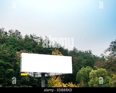 Blank white billboard mockup standing near a mountain green forest under clear sky, ideal for outdoor advertising, promotional campaigns, and design p Stock Photo