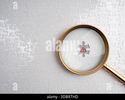 Magnifying glass highlighting red warning symbol on white missing puzzle piece, representing detection of scam, malware, or hidden threats in cybersec Stock Photo