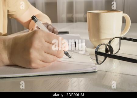 Person's hand writing in spiral notebook, with glasses and coffee mug nearby Stock Photo