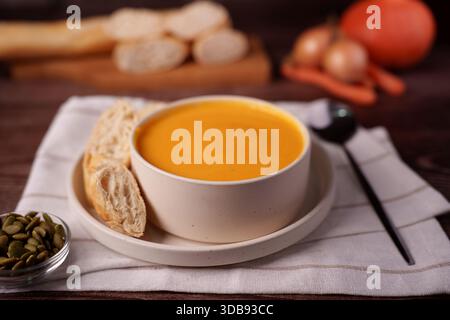 creamy pumpkin soup in white bowl with sliced bread and pumpkin seeds on linen napkin, warm rustic setting with onions and carrots in background, invi Stock Photo