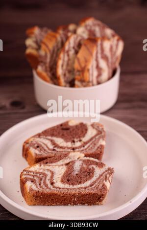 close-up view of sliced marble cake displayed on a white plate with additional slices in a bowl against a wooden background, highlighting the swirled Stock Photo