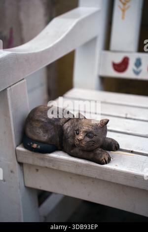 weathered wooden cat sculpture resting on rustic painted chair in a quiet outdoor setting with subtle weathered texture and earthy tones reflecting tr Stock Photo
