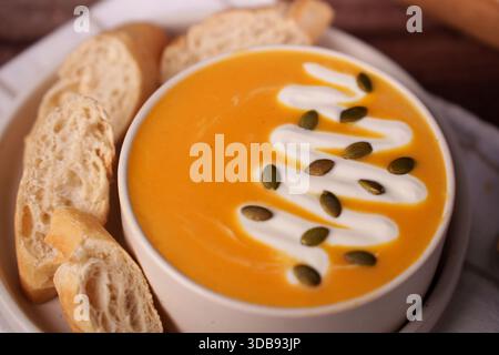 creamy pumpkin soup with whipped cream and pumpkin seeds in white bowl with slices of crisp baguette on the side creating a warm and inviting culinary Stock Photo