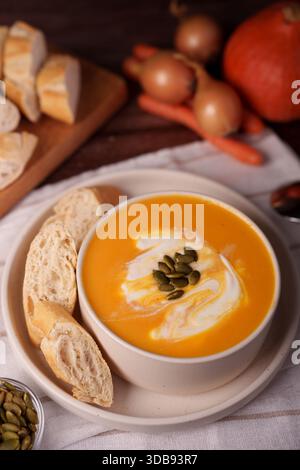 creamy pumpkin soup garnished with pepitas and cream swirls, served with freshly baked french baguette on rustic tablecloth, surrounded by autumn vege Stock Photo
