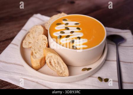 a cozy autumnal serving of creamy pumpkin soup garnished with cream and seeds, paired with fresh crusty bread, set on a rustic wooden table for a warm Stock Photo