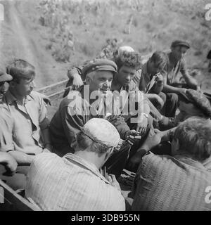 1970s. USSR. Donetsk region. A group of Soviet workers riding in the open back of a truck during the construction of strategic high-voltage 750 kV power lines. An older foreman in a cap tells a story while younger men in work clothes listen and smoke during a transport to the remote site. An authentic documentary shot vividly capturing the atmosphere of male camaraderie, collective labor spirit and daily life in the peaceful Donbas steppe before the war. Stock Photo