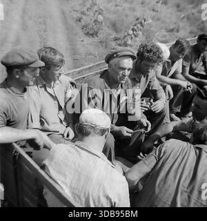 1970s. USSR. Donetsk region. A group of Soviet workers riding in the open back of a truck during the construction of strategic high-voltage 750 kV power lines. An older foreman in a cap tells a story while younger men in work clothes listen and smoke during a transport to the remote site. An authentic documentary shot vividly capturing the atmosphere of male camaraderie, collective labor spirit and daily life in the peaceful Donbas steppe before the war. Stock Photo