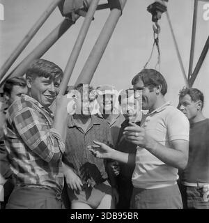 1970s. USSR. Donetsk region. Construction of strategic high-voltage 750 kV power lines. A group of young installers and engineers taking a break on the construction site. The men are standing near massive steel structures and a crane hook, dressed in summer clothes due to the heat. They are smoking cigarettes, laughing and having a lively conversation. A documentary shot capturing the atmosphere of male friendship and the working rhythm of energy builders in the peaceful Donbas steppe before the war. Stock Photo