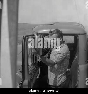 1970s. USSR. Donetsk region. Construction of strategic high-voltage 750 kV power lines. A charismatic portrait of a Soviet truck driver standing by the open door of his vehicle in the middle of the steppe. The man in a plaid shirt gestures expressively while talking to a colleague inside the cab or checking the route. A historic documentary shot capturing the daily routine of transport logistics and the atmosphere of labor in the peaceful Donbas before the war. Stock Photo