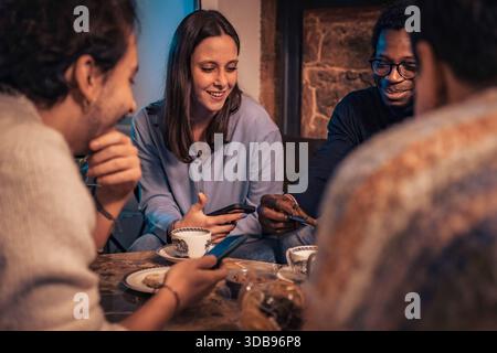A diverse group of young friends gather in a cozy, dimly lit cafe, sharing smiles and interacting with their smartphones over coffee during a casual e Stock Photo