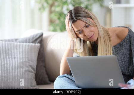 Worried woman is checking bad news on laptop sitting on a couch at home Stock Photo