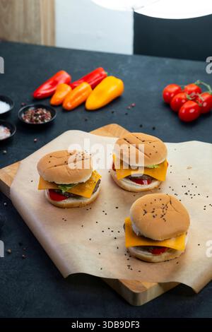Small cheeseburgers on parchment paper with various fresh vegetables and spices on a dark background Stock Photo