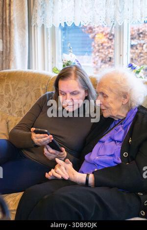 Adult daughter showing pictures on smartphone to her elderly mother sitting on sofa in living room Stock Photo