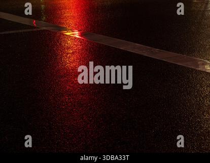 Wet asphalt reflecting vibrant red and yellow lights at night, shimmering around a white road marking to create an abstract urban texture and moody ra Stock Photo