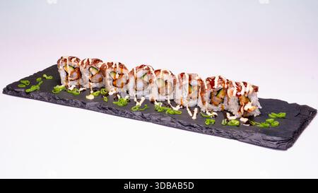 Uramaki sushi rolls with fried filling, avocado and cucumber, drizzled with sauce and garnished with spring onion on slate board. Stock Photo