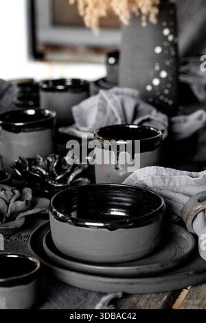 Rustic table setting with empty craft handmade ceramic tableware, black bowls, plates, cups, jug and vase on linen tablecloth. Blck ceramic roses flow Stock Photo