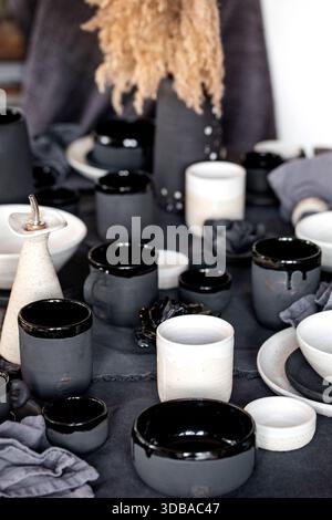 Rustic table setting with empty craft handmade ceramic tableware, black and white rough bowls, cups, jug and vase on linen tablecloth. Dry reed flower Stock Photo