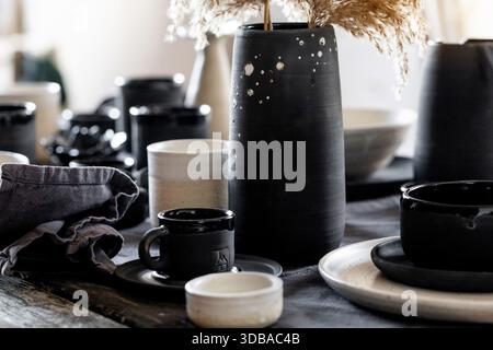 Rustic table setting with empty craft handmade ceramic tableware, black and white rough bowls, cups, jug and vase on linen tablecloth. Dry reed flower Stock Photo