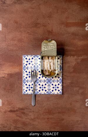 Open tin of sardine fillets in oil, served with fork on white-and-blue patterned ceramic tiles. Textured brown background, top view. Colors of the yea Stock Photo