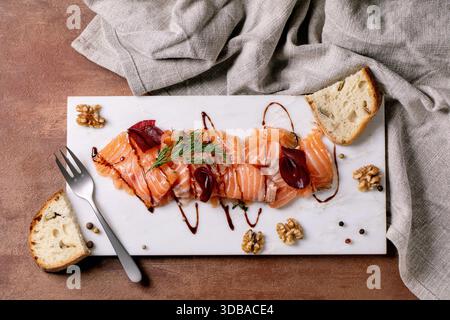 Thinly sliced salted salmon fillet served with beetroot slices, walnuts, balsamic vinegar, and fresh bread on a white marble board, fine dining. Brown Stock Photo