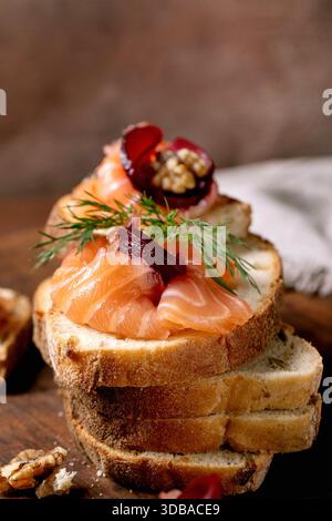 Sandwiches with thinly sliced salted salmon fillet, beetroot slices, walnuts, balsamic vinegar and fresh wheat bread in stack on wooden board. Brown b Stock Photo