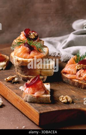 Sandwiches with thinly sliced salted salmon fillet, beetroot slices, walnuts, balsamic vinegar and fresh wheat bread in stack on wooden board. Brown b Stock Photo