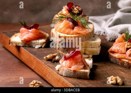 Sandwiches with thinly sliced salted salmon fillet, beetroot slices, walnuts, balsamic vinegar and fresh wheat bread in stack on wooden board. Brown b Stock Photo