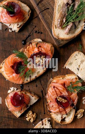 Sandwiches with thinly sliced salted salmon fillet, sardines, beetroot slices, walnuts, balsamic vinegar and fresh wheat bread on wooden board. Brown Stock Photo