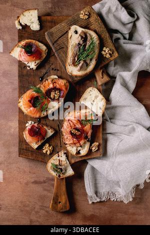 Sandwiches with thinly sliced salted salmon fillet, sardines, beetroot slices, walnuts, balsamic vinegar and fresh wheat bread on wooden board. Brown Stock Photo
