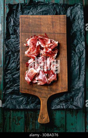 Thinly sliced Spanish ham Jamon Iberico paleta on wooden cutting board. Old dark turquoise wooden background, crumpled black paper, top view. Stock Photo